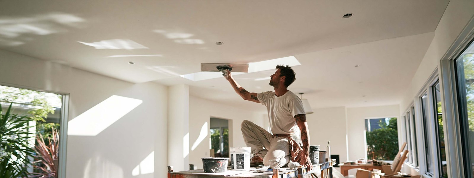 A drywaller working across a wide ceiling in daylight, buckets and tools on the plank beside him
