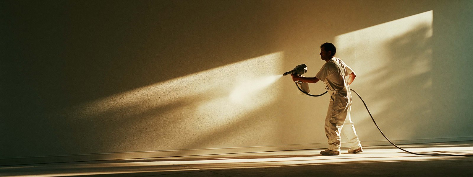 A plasterer working a spray gun along a wide wall, light raking across the finished surface