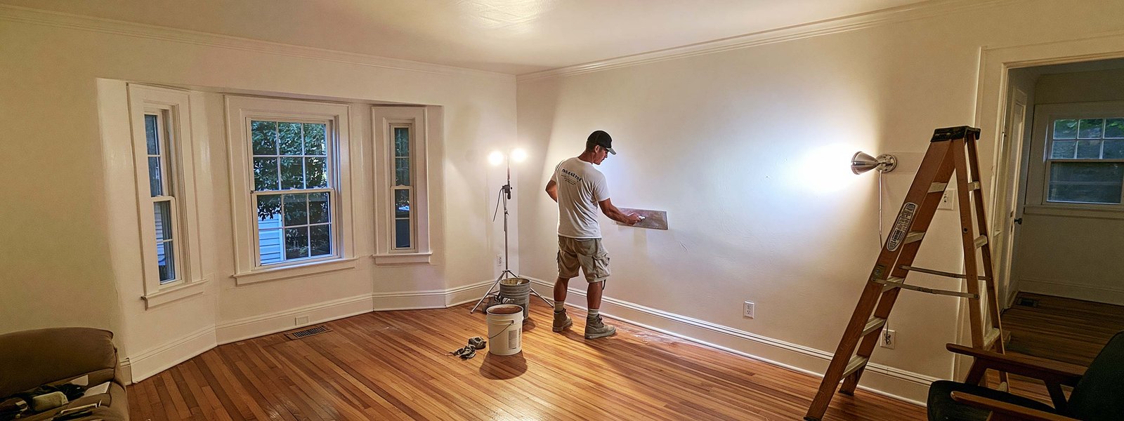 A plasterer working along a wall in a pre-war room with a bay window and wood floors