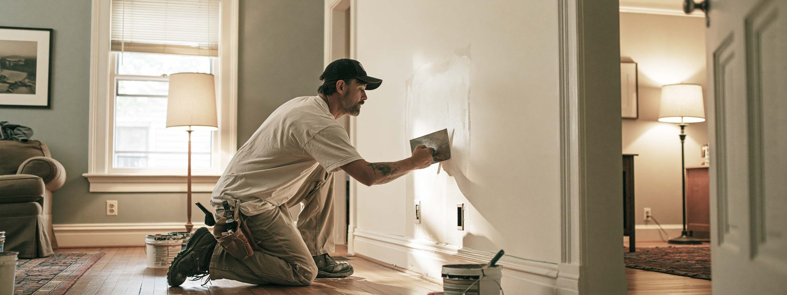 A drywaller working on a small repair in a lived-in room, hardwood floor and furniture around him