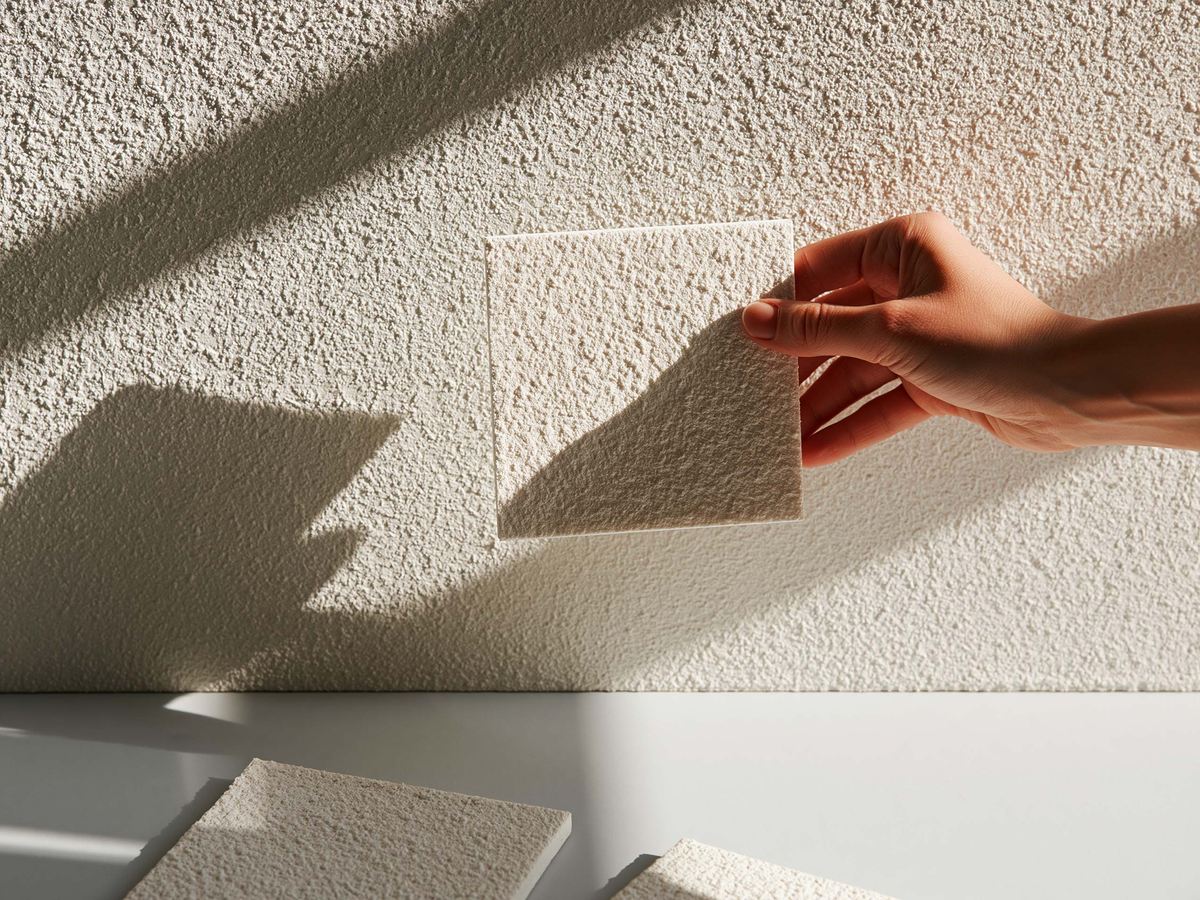A hand holding a sample board against a textured wall to compare the two finishes