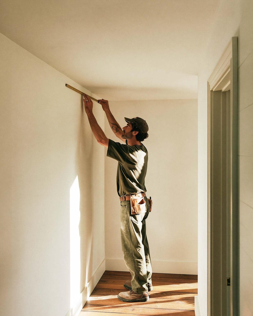 A drywaller reaching up to measure a wall near the ceiling in an empty room, afternoon sun across the boards