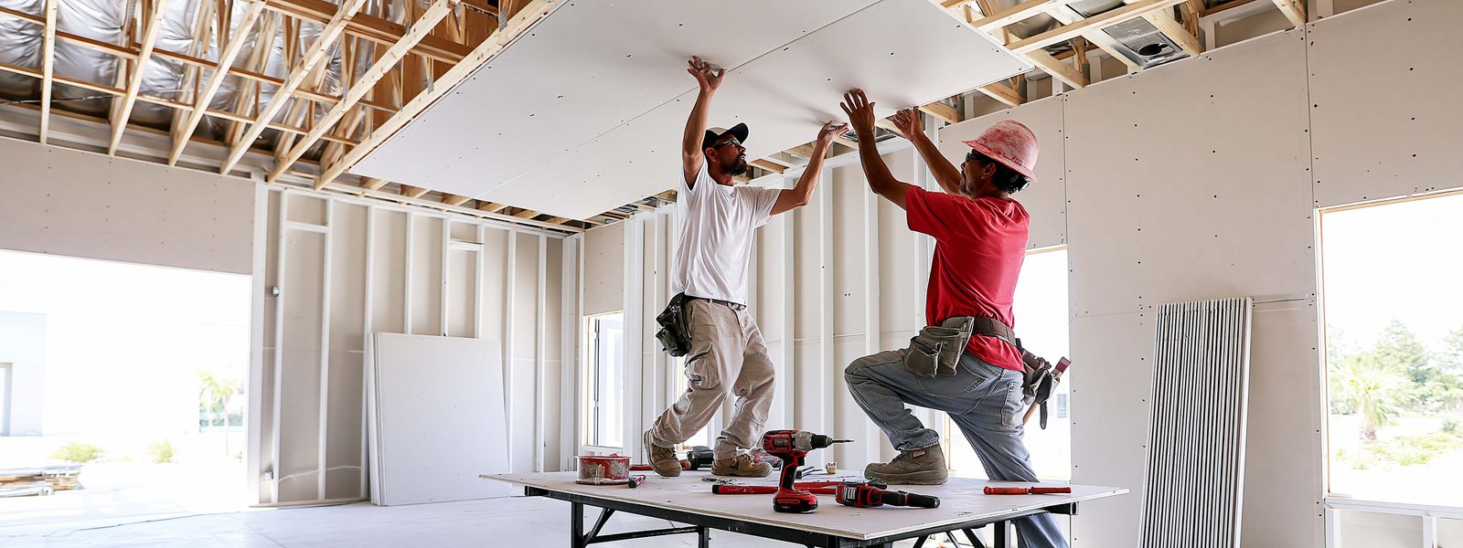 Two drywallers lifting a full sheet into place against ceiling joists in an unfinished room