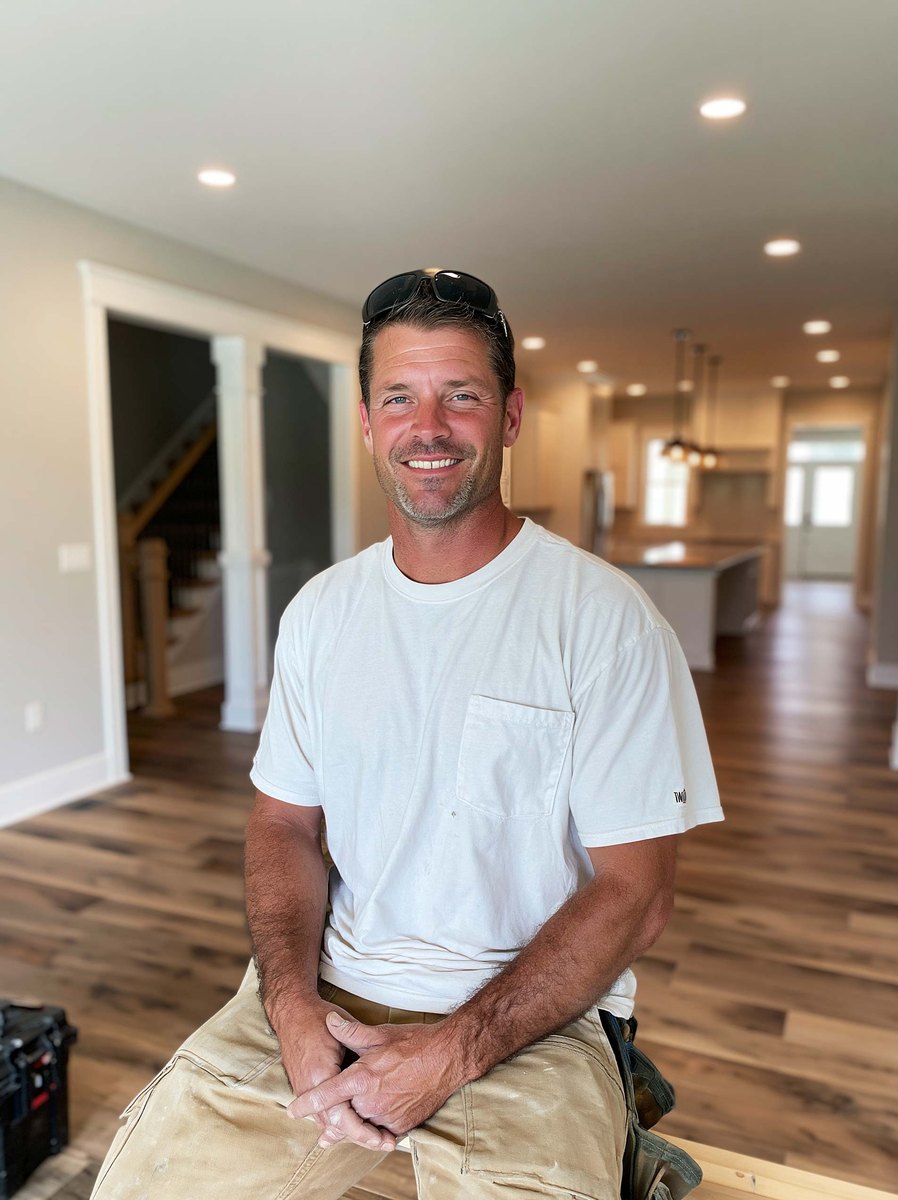 Drywall contractor in a white t-shirt sitting on a plank in a newly finished home, kitchen and staircase behind him